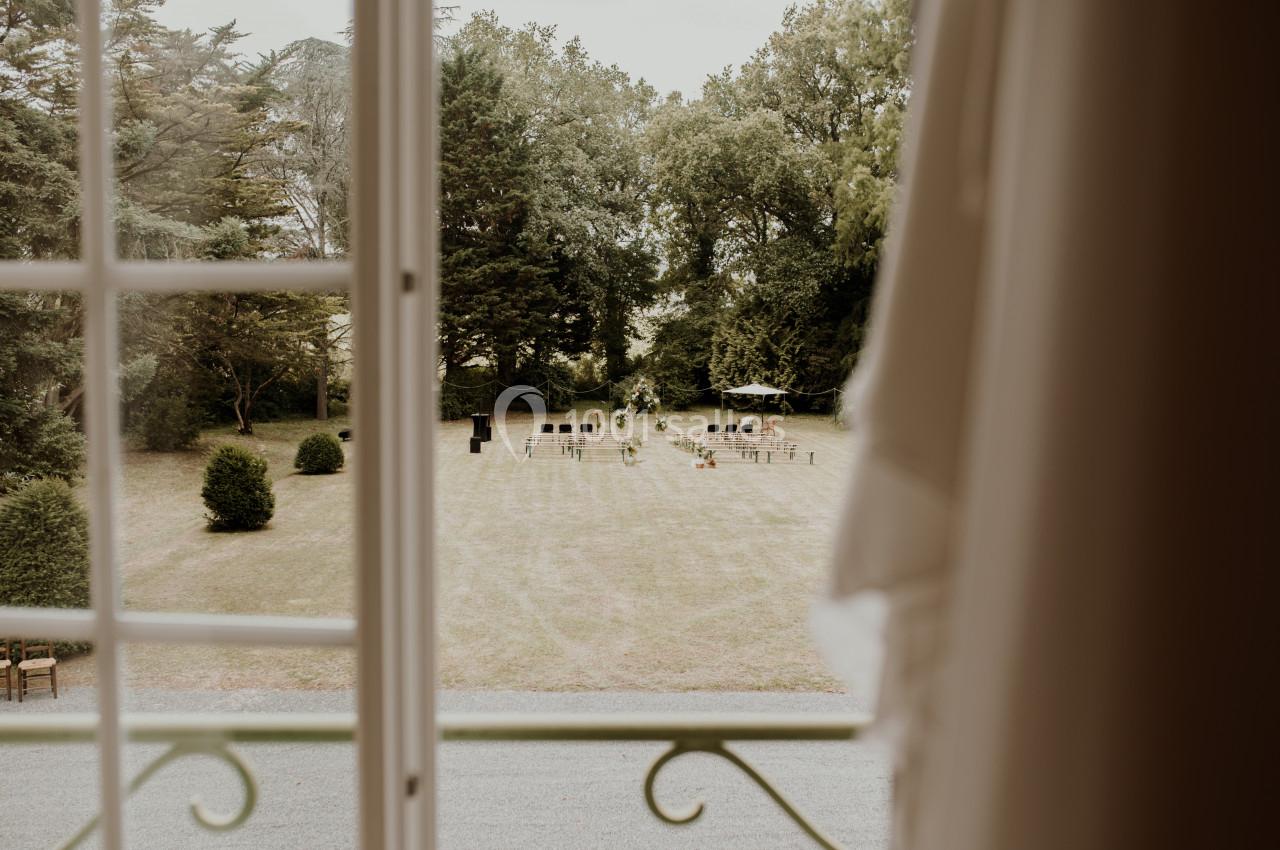 Vue depuis une fenêtre ouverte sur un jardin avec des chaises disposées pour un événement en plein air.
