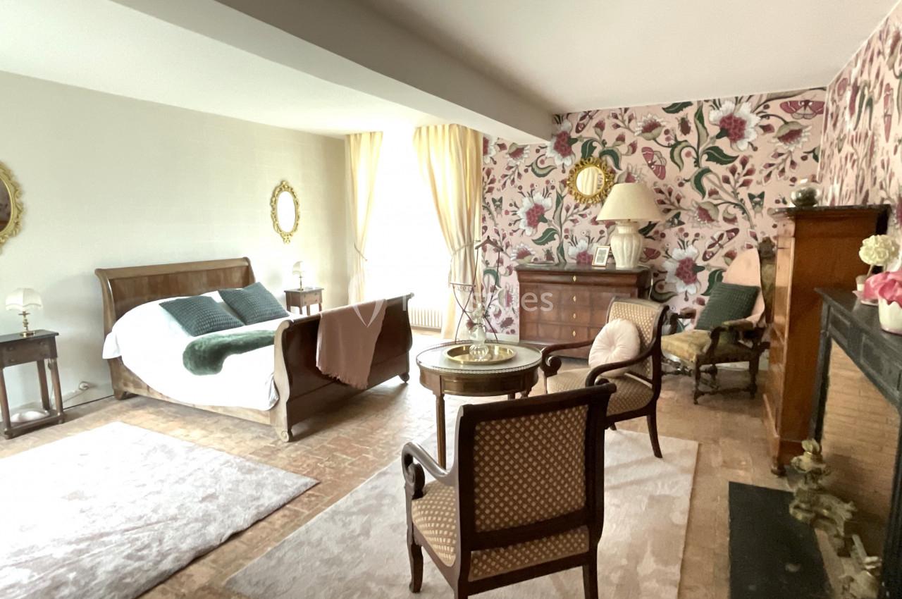 Chambre lumineuse avec lit double, fauteuils, cheminée, meubles anciens et mur décoré de motifs floraux.