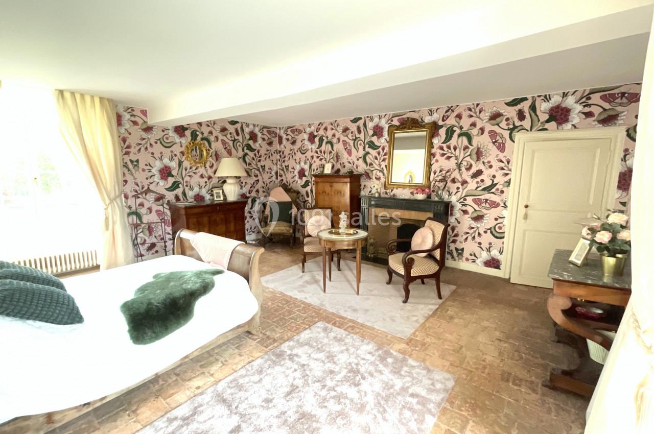 Chambre lumineuse avec lit, fauteuils, cheminée, meubles en bois et murs décorés de papier peint floral.