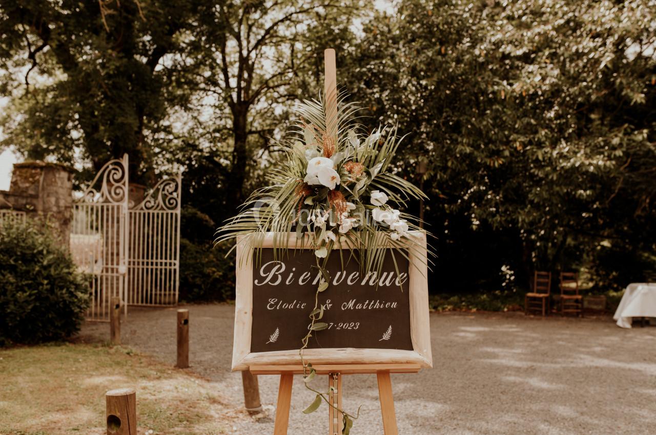 Panneau de bienvenue décoré de fleurs, placé sur un chevalet dans un jardin avec portail en arrière-plan.