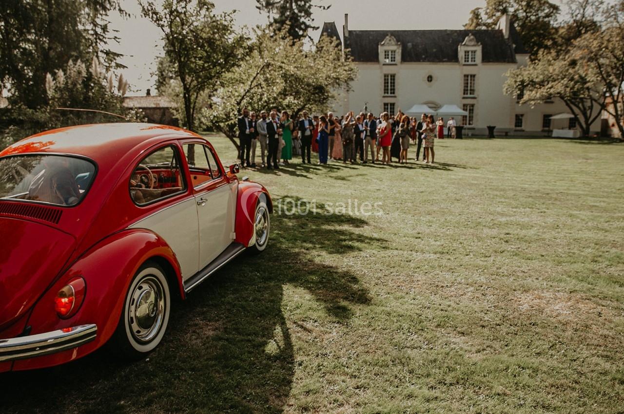 Une voiture rouge est garée sur une pelouse devant un groupe de personnes rassemblées près d'un bâtiment ancien.