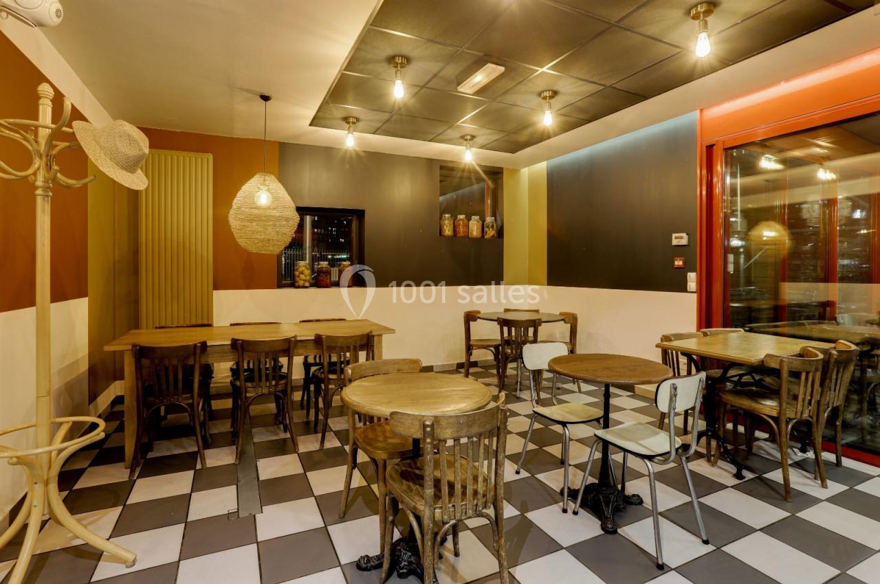 Salle de restaurant avec tables en bois, chaises dépareillées, éclairage suspendu et sol à carreaux noir et blanc.