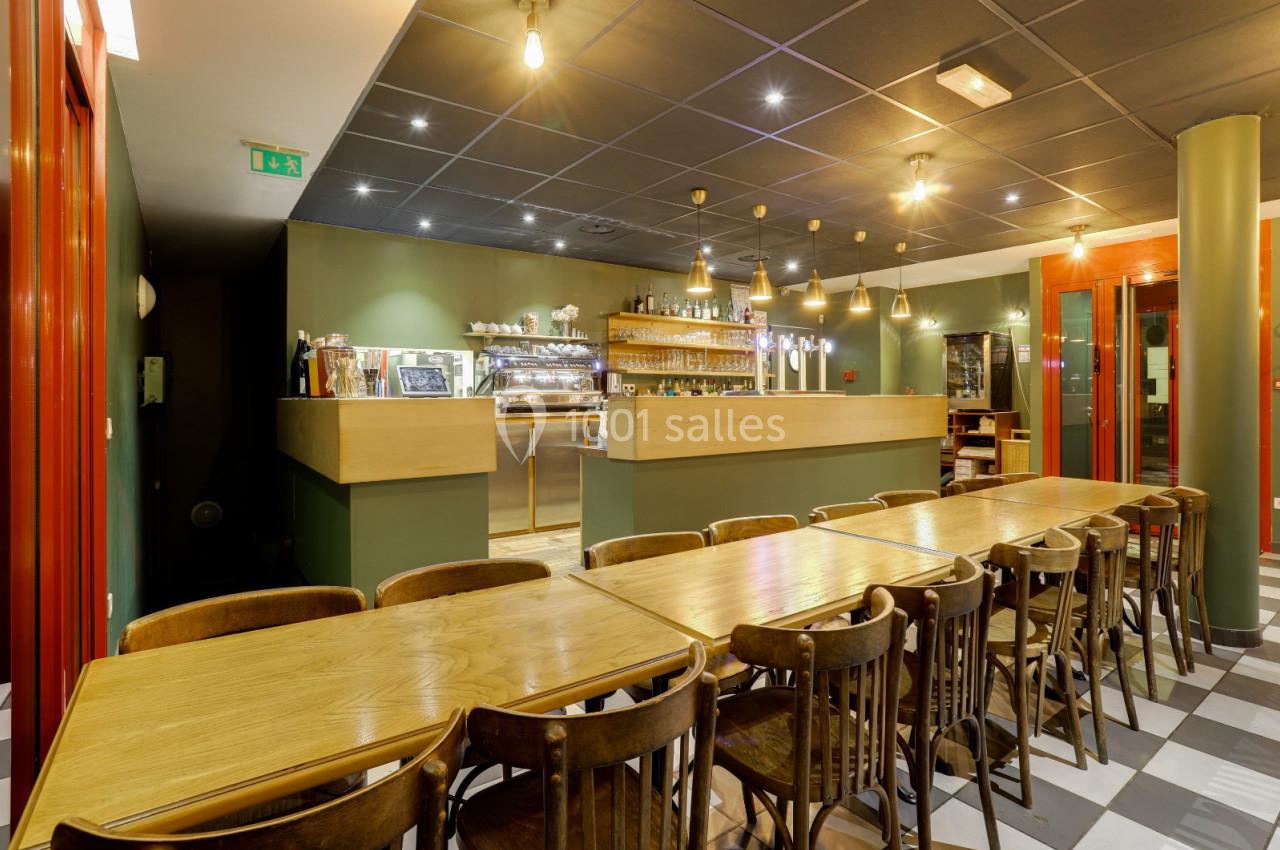 Salle de restaurant avec longues tables en bois, chaises assorties et comptoir de bar éclairé en arrière-plan.