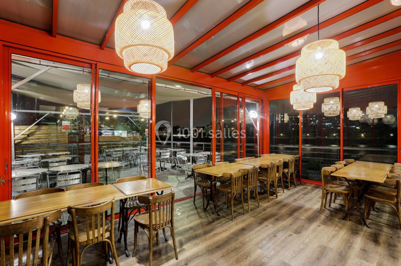 Salle de restaurant avec tables en bois, chaises assorties et grandes lampes suspendues, donnant sur une terrasse vitrée.