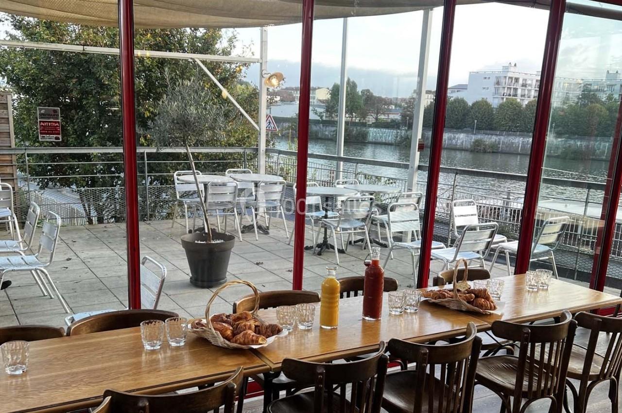 Salle de restaurant avec une grande table en bois, vue sur une terrasse extérieure et un cours d'eau en arrière-plan.