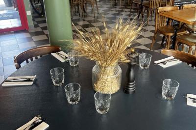 Table ronde dressée avec des verres, couverts et un centre de table en épis de blé dans un restaurant au sol carrelé.