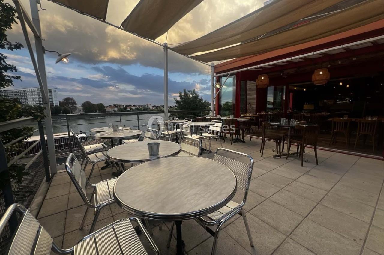 Terrasse d'un café avec des tables et chaises métalliques, vue sur un plan d'eau et un ciel partiellement nuageux.