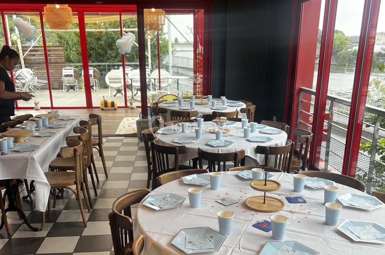 Salle décorée pour un événement avec tables dressées, nappes blanches, vaisselle bleue et vue sur une terrasse extérieure.