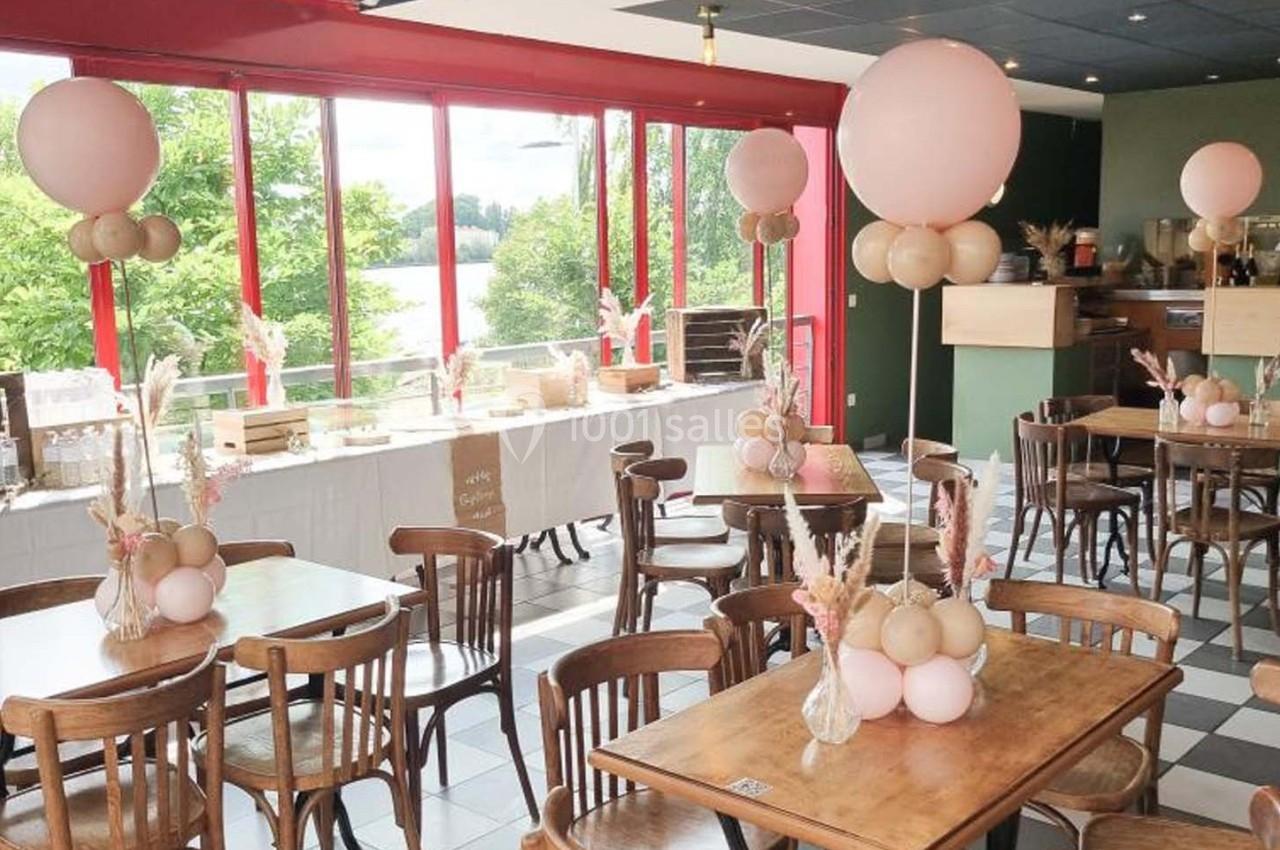 Salle décorée pour un événement avec des tables en bois, des ballons roses et une vue sur un paysage extérieur verdoyant.