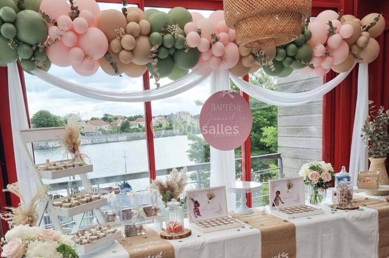 Décoration de baptême avec ballons pastel, table garnie de douceurs et vue sur un paysage extérieur avec rivière.