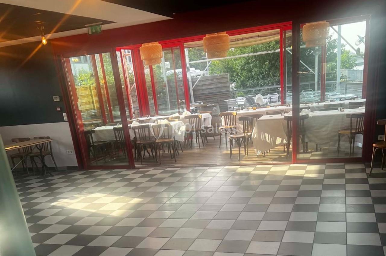 Salle de restaurant lumineuse avec sol en damier, tables dressées derrière une baie vitrée donnant sur une terrasse.