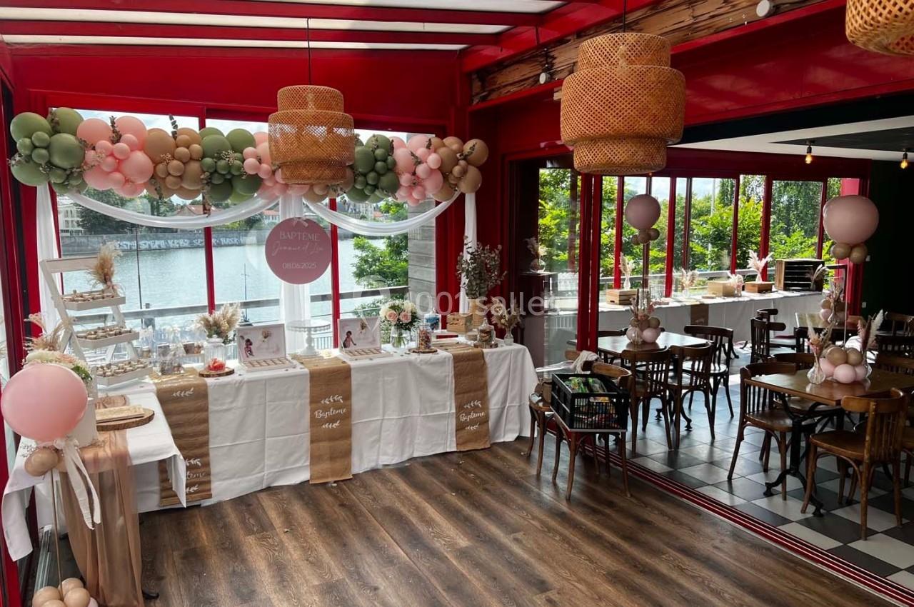 Salle décorée pour une fête avec ballons, nappes blanches, guirlandes et tables dressées près de grandes fenêtres.