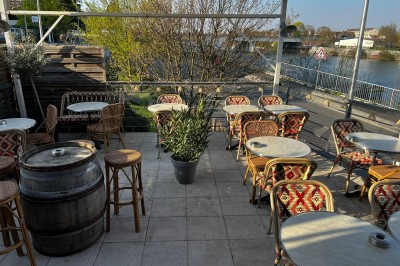 Terrasse extérieure avec tables, chaises en osier, tonneau en bois et vue sur un fleuve bordé de végétation.