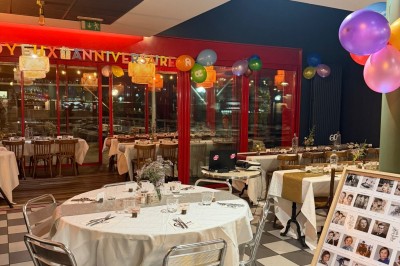 Terrasse extérieure avec tables, chaises en osier, tonneau en bois et vue sur un fleuve bordé de végétation.