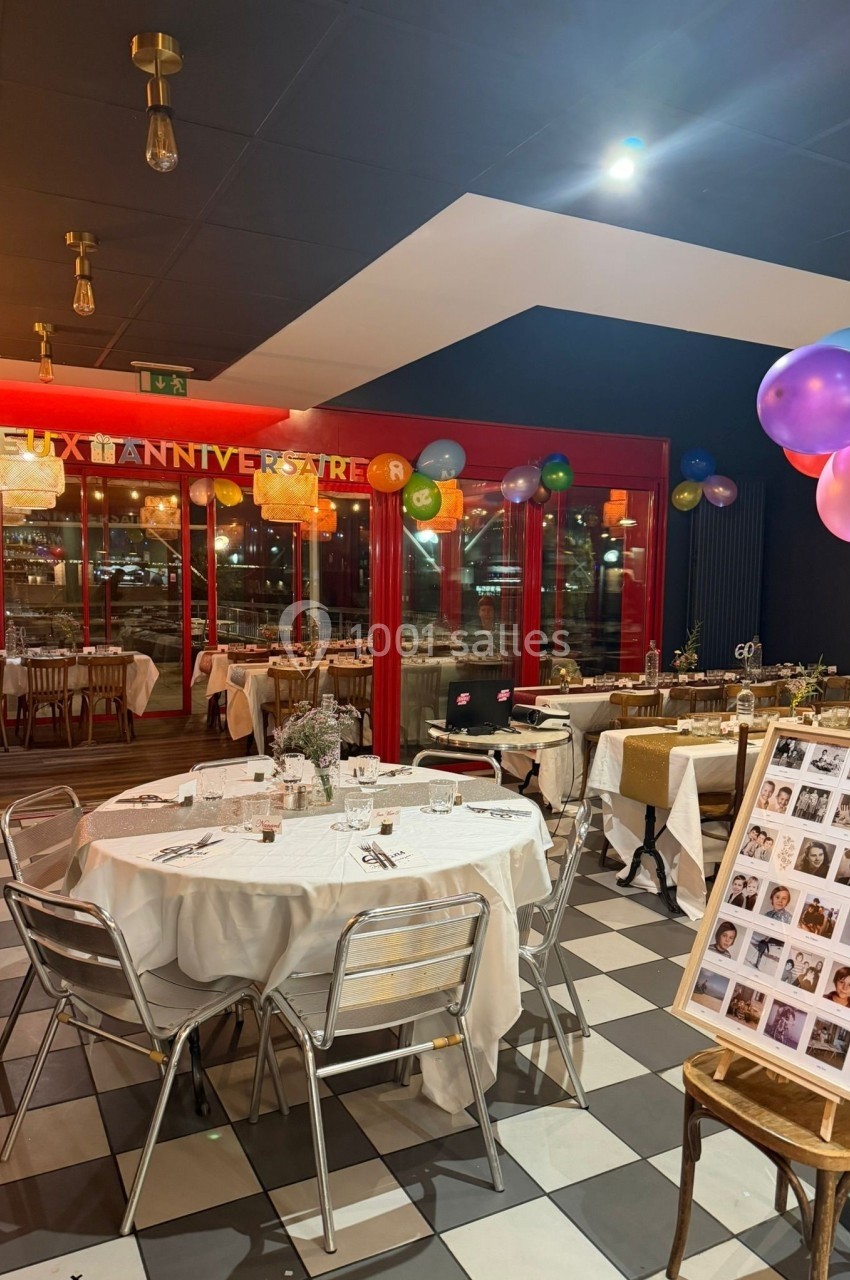 Salle décorée pour une fête avec tables dressées, ballons colorés et un panneau de photos près de l'entrée.