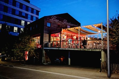 Terrasse extérieure avec tables, chaises en osier, tonneau en bois et vue sur un fleuve bordé de végétation.