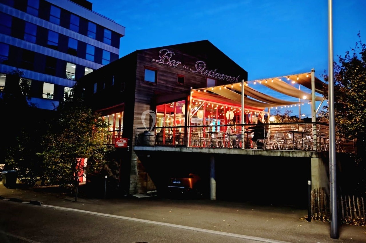 Terrasse d'un bar-restaurant éclairée en soirée, avec des tables et chaises sous une pergola lumineuse.