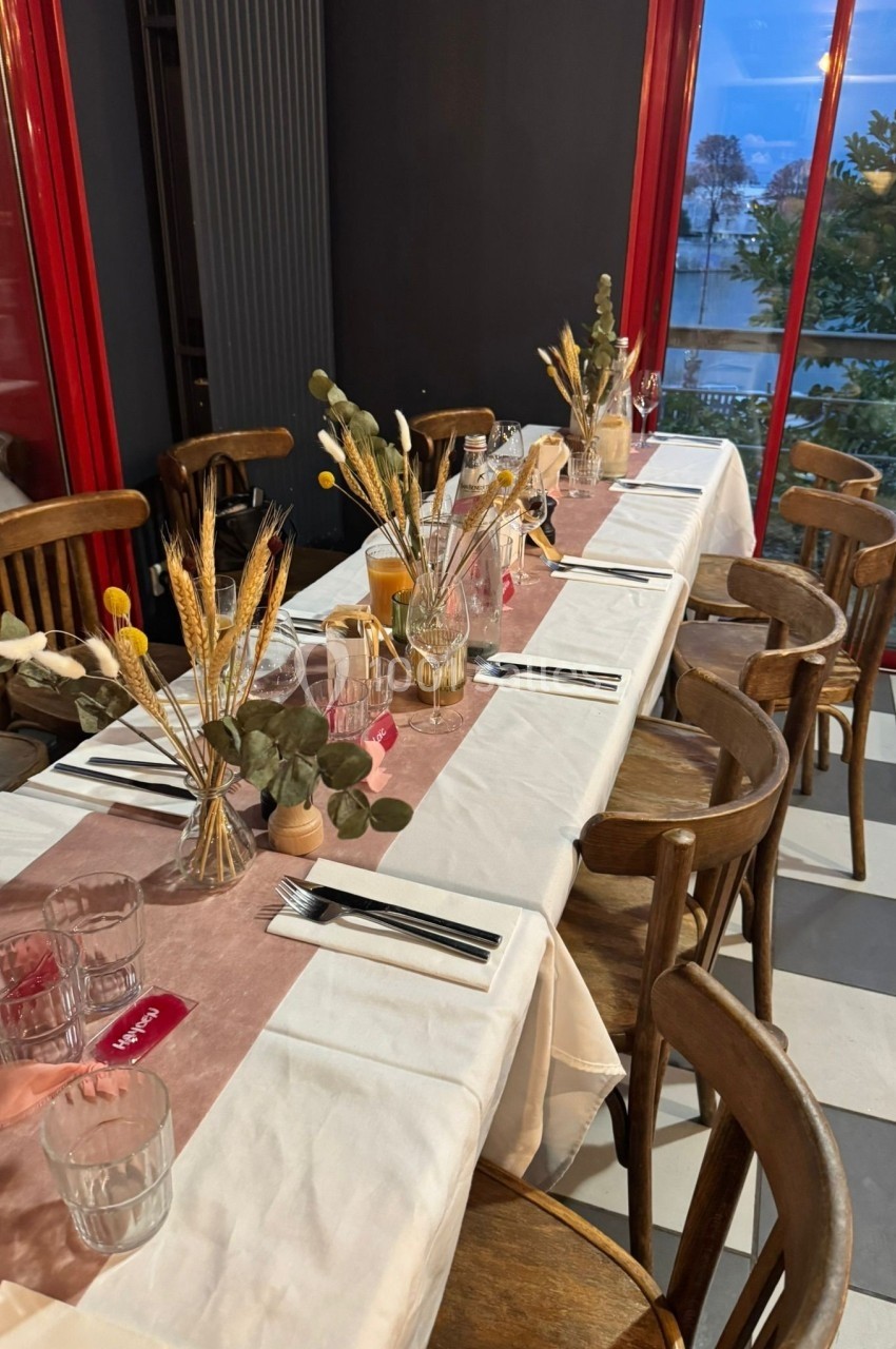 Table dressée avec nappe blanche, chemin de table rose, décorations florales et couverts dans une salle lumineuse.