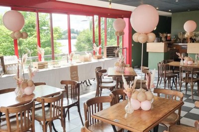 Terrasse extérieure avec tables, chaises en osier, tonneau en bois et vue sur un fleuve bordé de végétation.