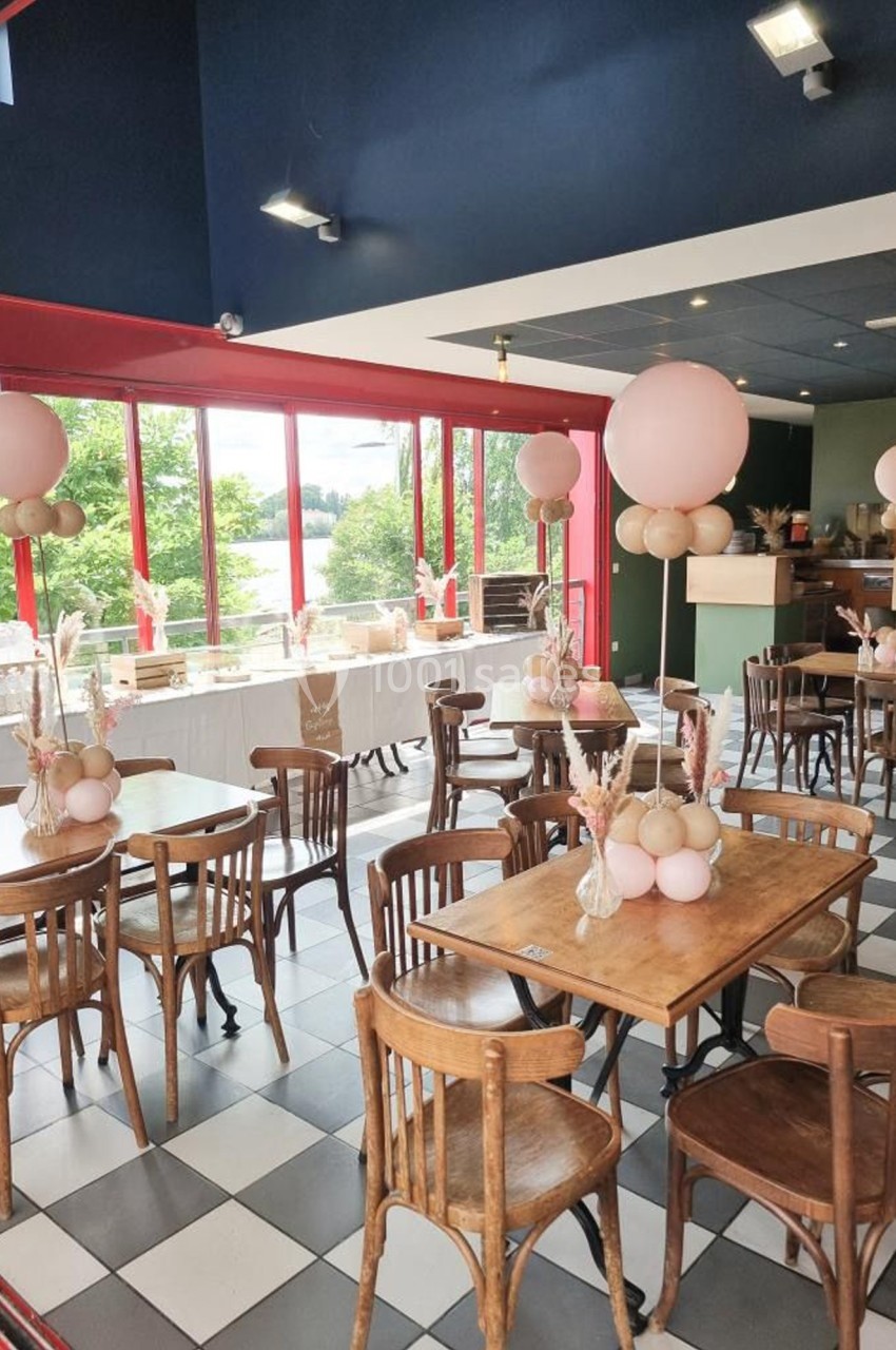 Salle de restaurant lumineuse avec tables en bois décorées de ballons roses et vue sur un espace extérieur verdoyant.