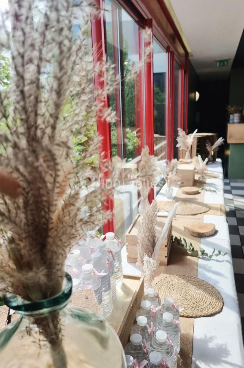 Table décorée avec des vases de fleurs séchées, bouteilles d'eau et sets en osier, près d'une grande baie vitrée.