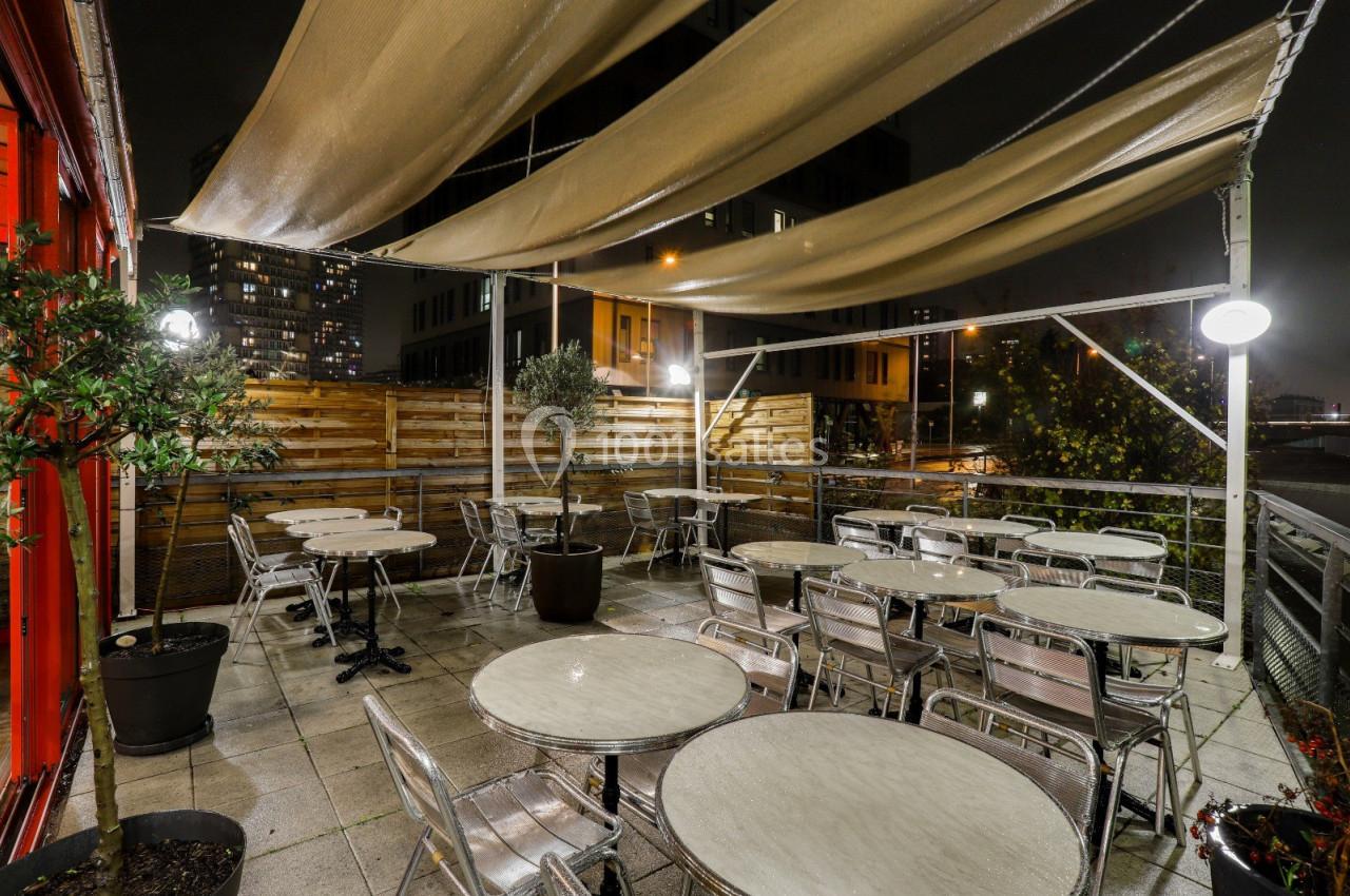 Terrasse extérieure avec tables et chaises en métal, éclairée la nuit, protégée par des toiles tendues.