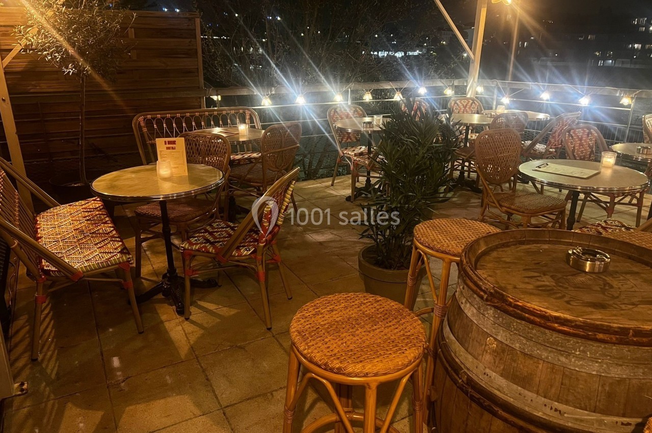 Terrasse extérieure éclairée en soirée avec tables, chaises en rotin, tonneaux en bois et plantes décoratives.