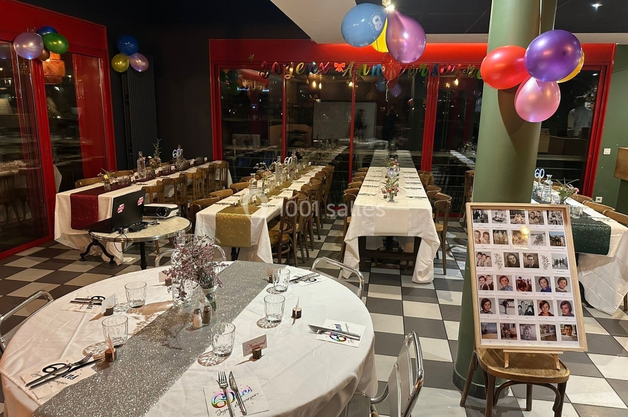 Salle décorée pour une fête avec tables dressées, ballons colorés et photos affichées sur une chaise.