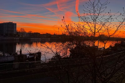 Coucher de soleil vibrant avec des nuages colorés se reflétant sur une rivière bordée d'arbres et de bâtiments.