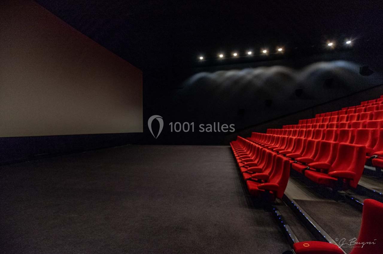 Salle de cinéma vide avec rangées de sièges rouges et un grand écran éteint dans une ambiance tamisée.