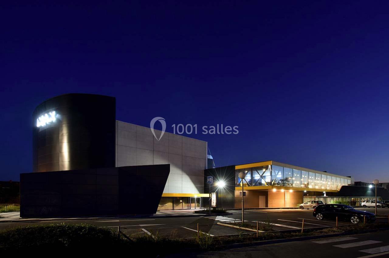 Bâtiment moderne éclairé de nuit avec un parking à l'avant, ciel sombre en arrière-plan.