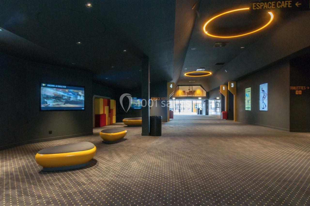 Couloir d'un cinéma avec sièges arrondis, écrans muraux et signalisation vers l'espace café et les toilettes.