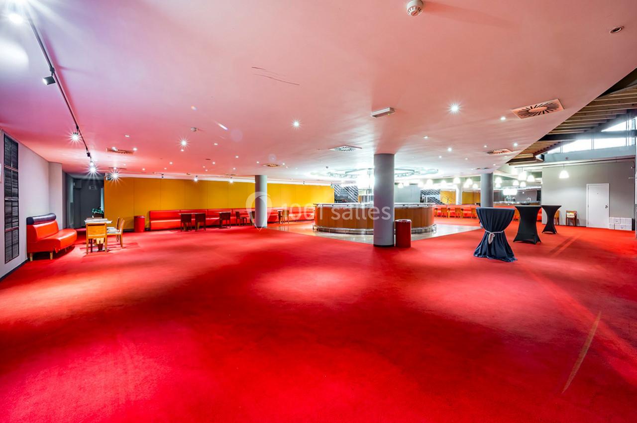 Salle spacieuse avec moquette rouge, mobilier moderne, tables hautes et éclairage lumineux.