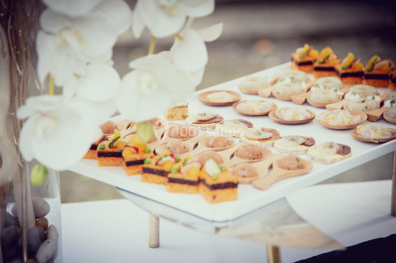Plateau de petites bouchées salées variées disposées sur des cuillères en bois, avec des fleurs blanches en avant-plan.