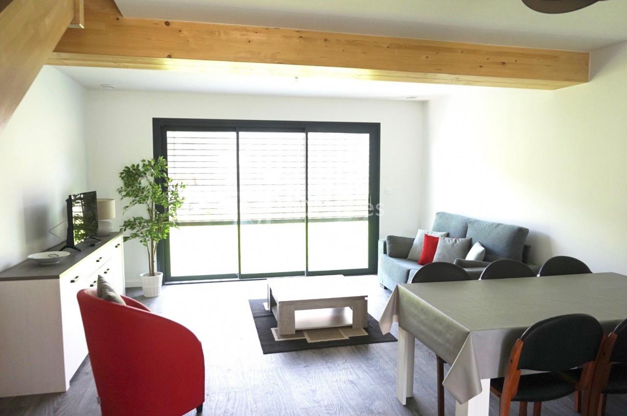 Salon lumineux avec canapé, table à manger, fauteuil rouge, meuble TV, plante verte et grande baie vitrée.