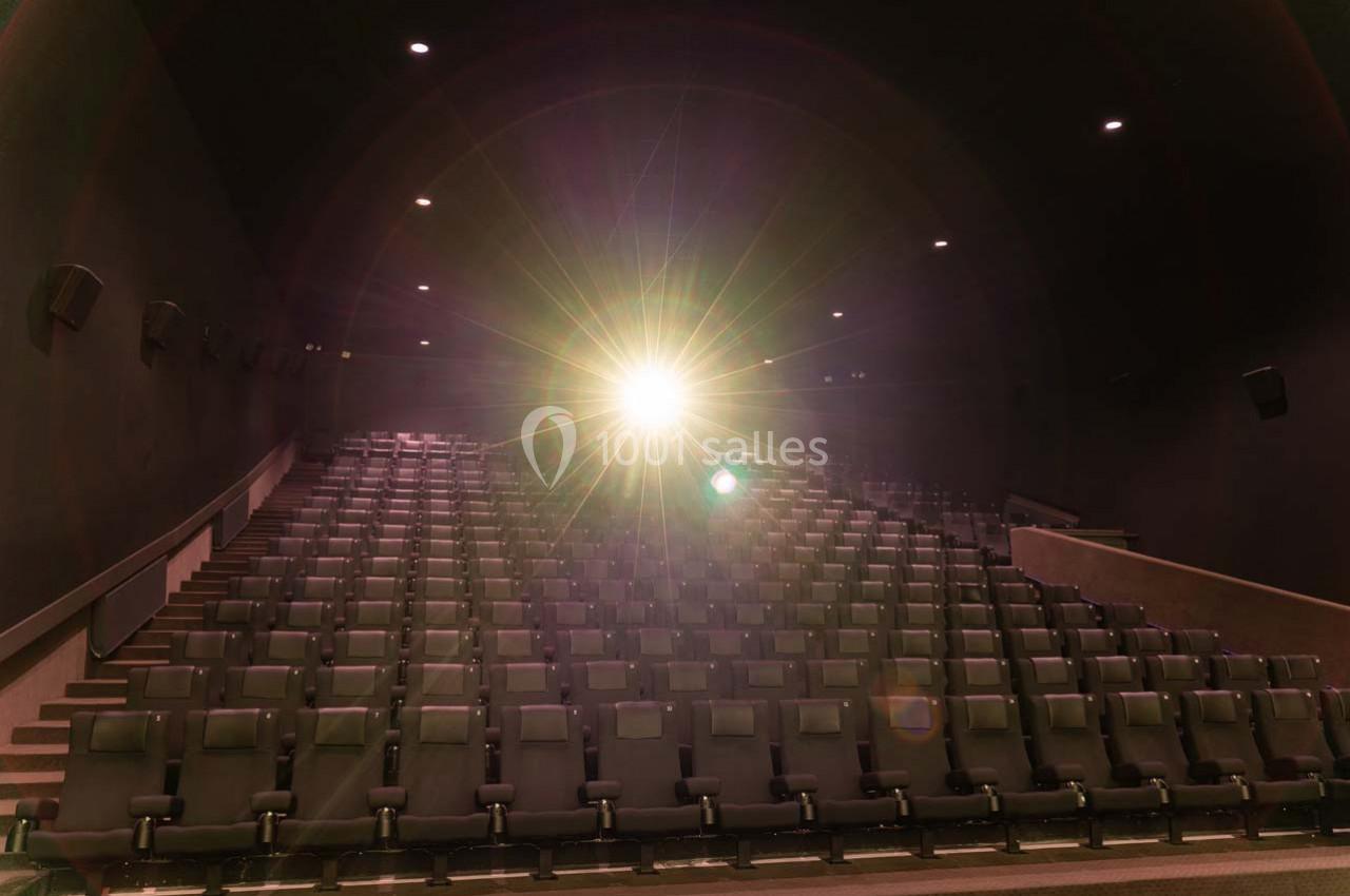 Salle de cinéma vide avec des rangées de sièges noirs et un projecteur allumé au centre.