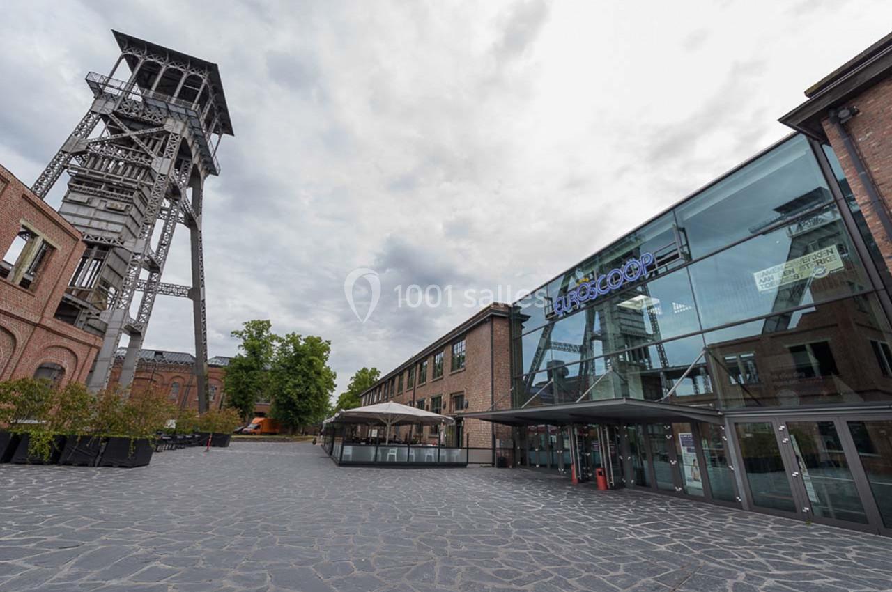 Vue d'un bâtiment moderne en verre à côté d'un ancien chevalement minier sous un ciel nuageux.