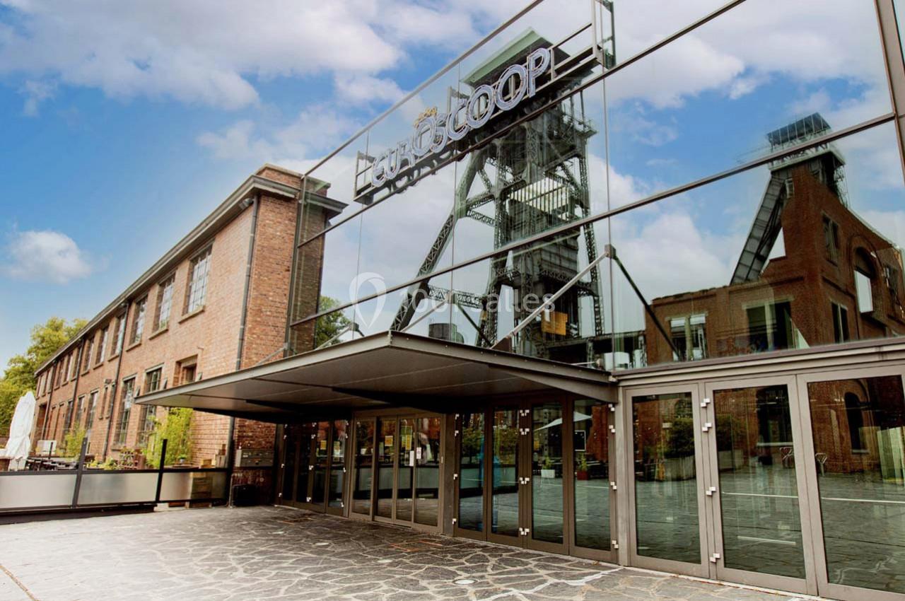 Façade vitrée d'un bâtiment industriel rénové avec une ancienne structure minière visible en arrière-plan.