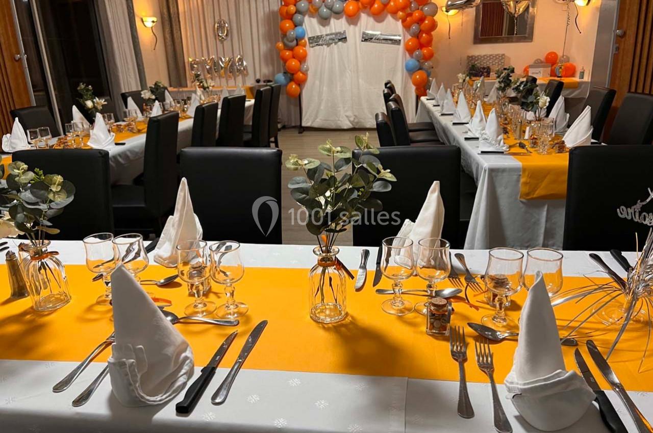 Salle décorée pour une réception avec tables dressées, chemins de table orange et ballons en arrière-plan.