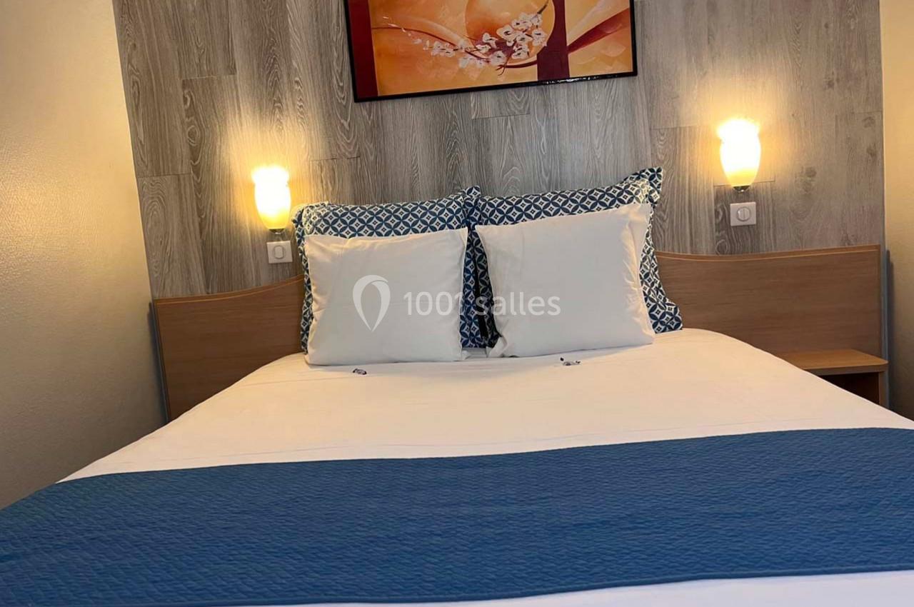 Lit double avec draps blancs, couverture bleue et coussins décoratifs, encadré par deux lampes murales allumées.