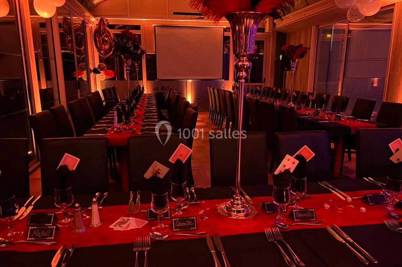 Salle décorée pour un événement, avec tables alignées, nappes rouges et noires, cartes à jouer et plumes en centre de table.