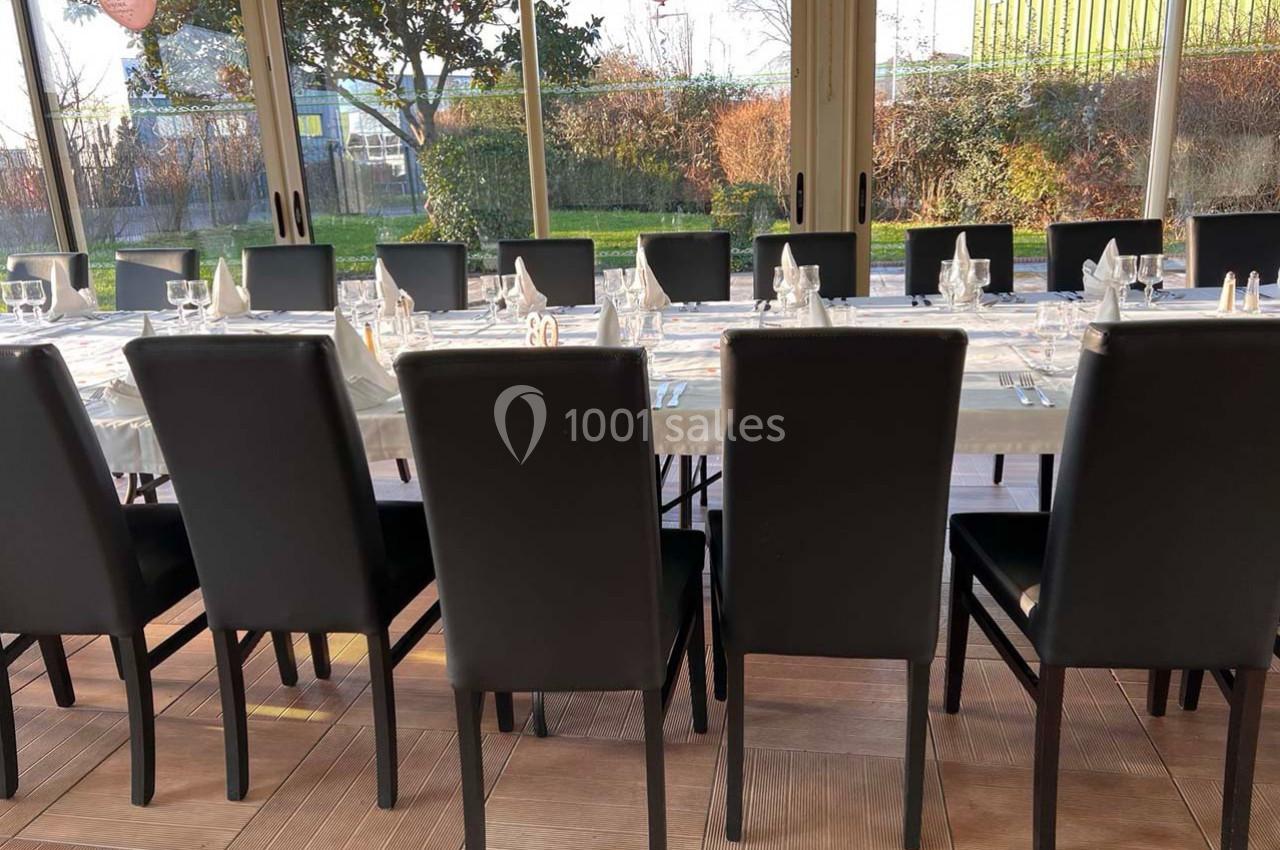 Salle avec des tables dressées pour un repas, entourées de chaises noires, et vue sur un jardin à travers des baies vitrées.