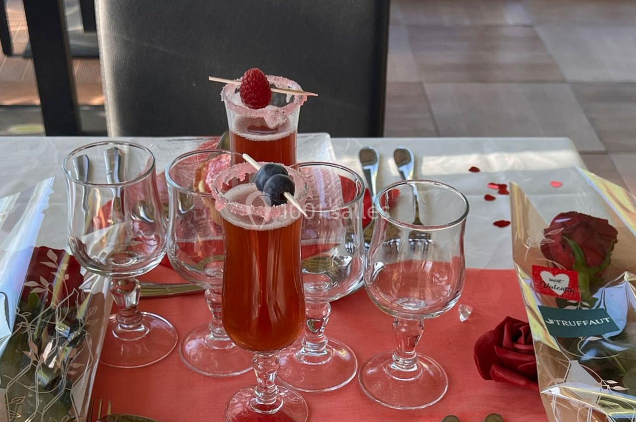 Verres de cocktail rouge décorés de fruits, entourés de verres à pied vides sur une table dressée avec une rose.