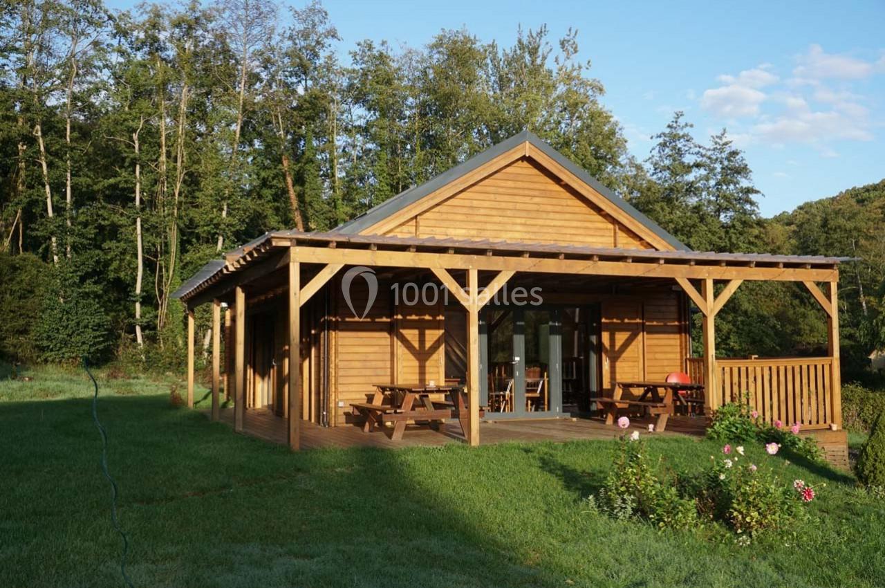 Chalet en bois avec terrasse couverte, entouré de pelouse et d'arbres, sous un ciel partiellement dégagé.