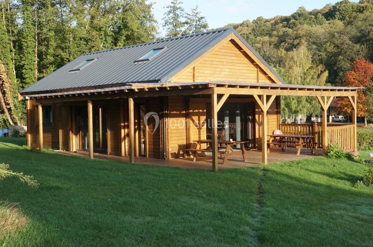 Maison en bois avec terrasse couverte, entourée de pelouse et d'arbres dans un environnement naturel.