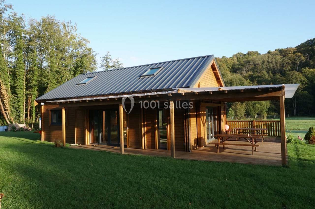 Maison en bois avec toit en métal, terrasse couverte et table de pique-nique, entourée de pelouse et d'arbres.