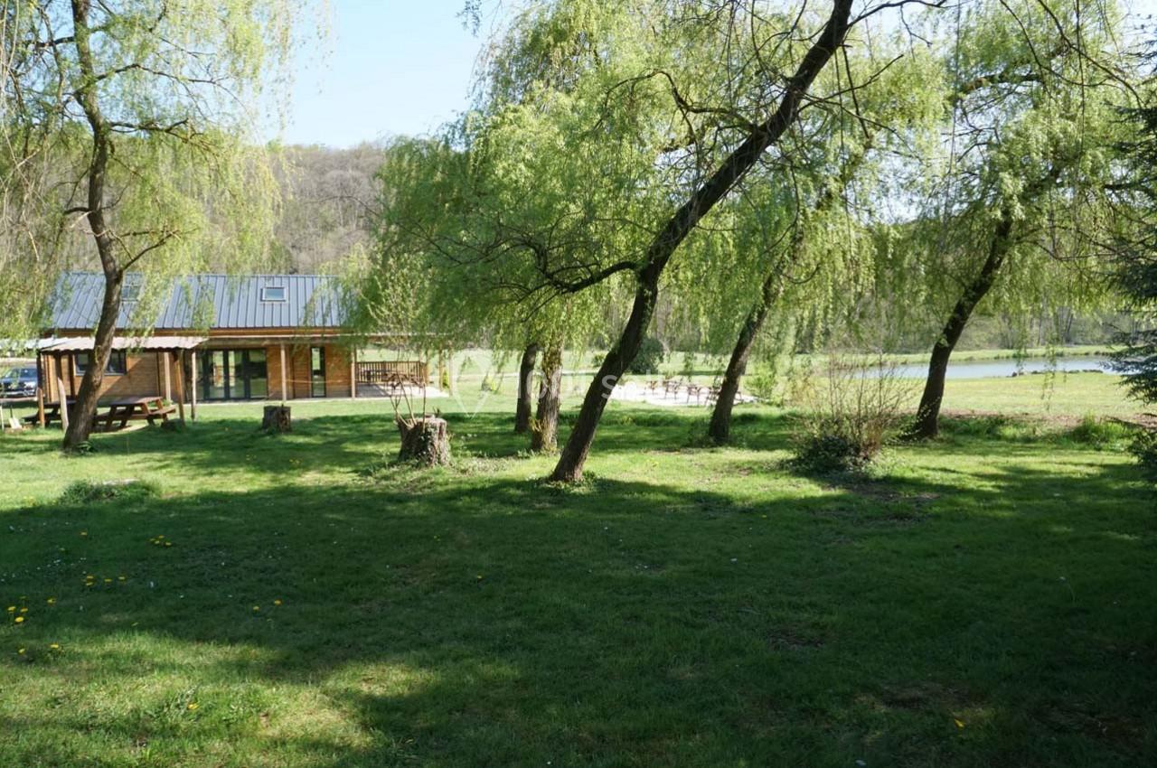 Maison en bois entourée d'arbres et d'une pelouse, située près d'un étang sous un ciel dégagé.