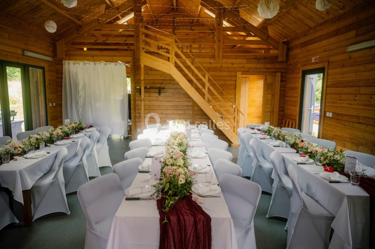 Salle en bois décorée pour un repas, avec deux longues tables dressées et ornées de fleurs.