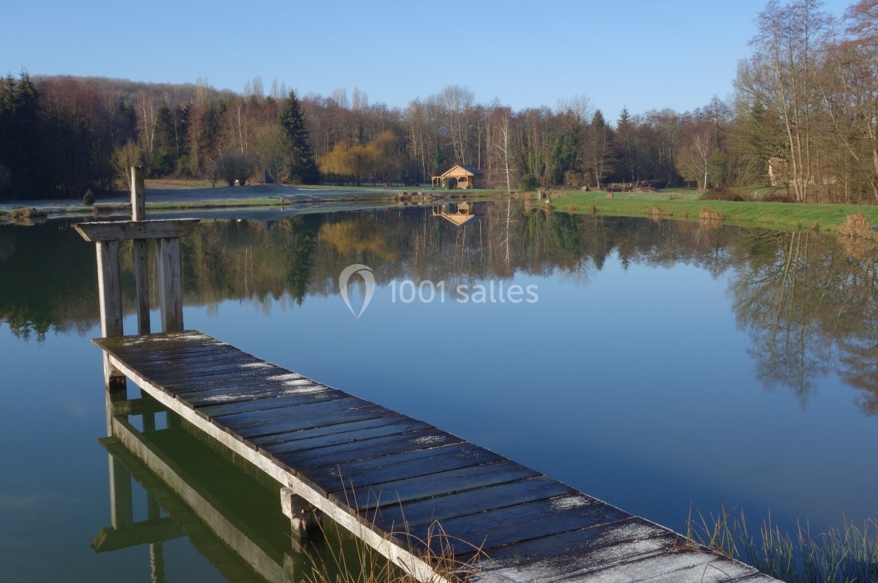 Ponton en bois au bord d’un lac calme entouré d’arbres et d’une cabane en arrière-plan sous un ciel dégagé.