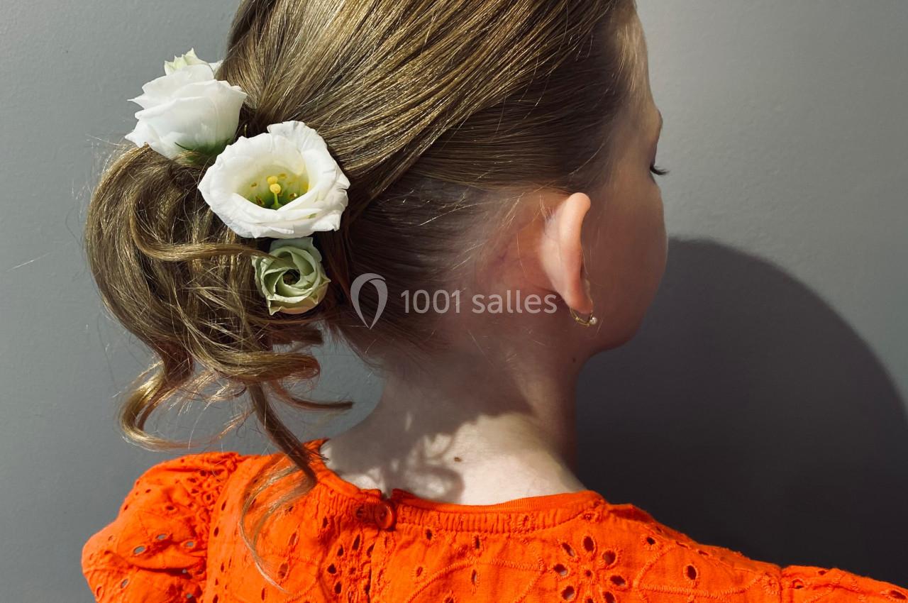 Une fillette de dos avec une coiffure relevée ornée de fleurs blanches, portant une robe orange à motifs brodés.